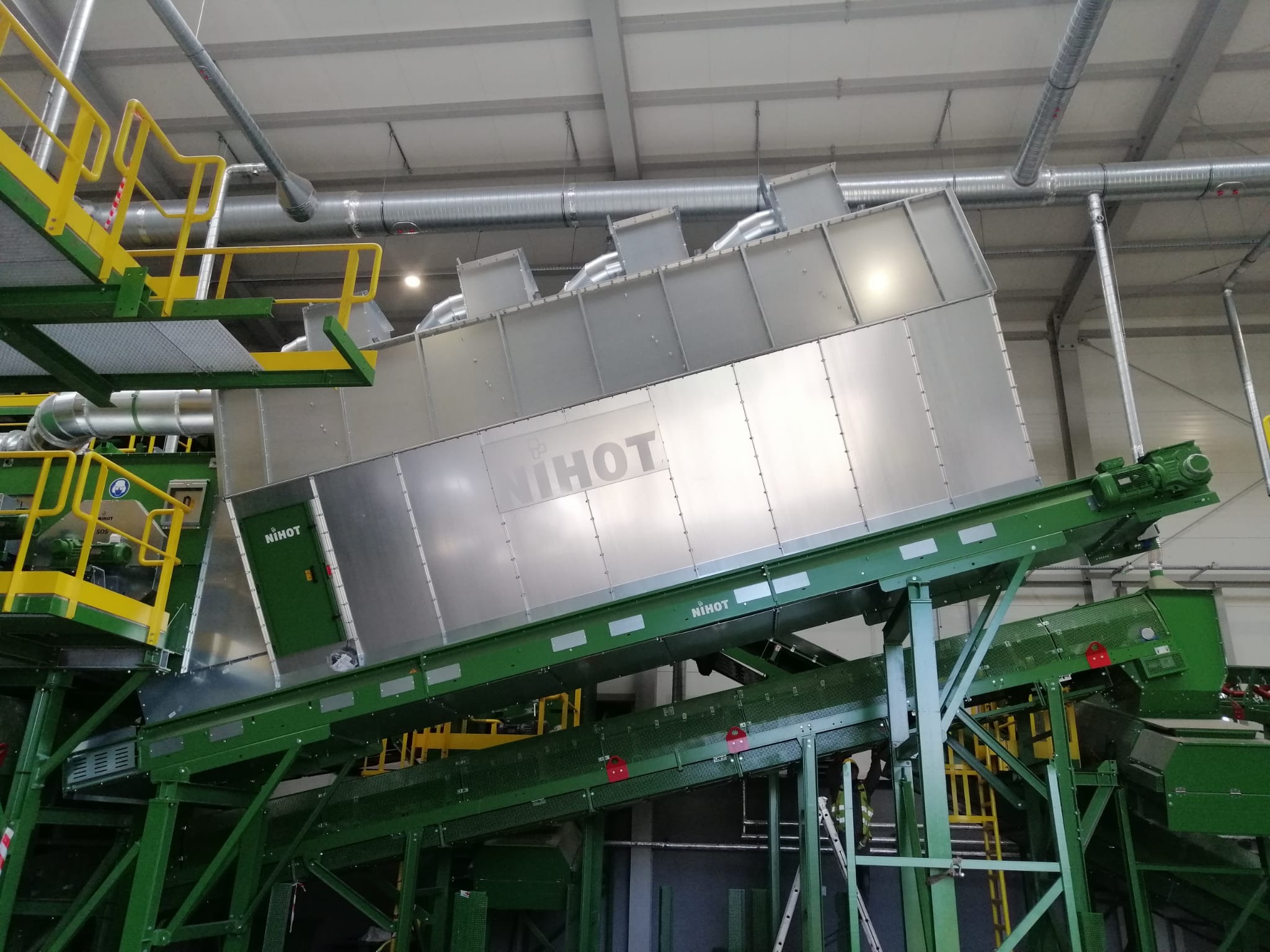 Wnętrze hali produkcyjnej z metalową konstrukcją sortowni z napisem NIHOT, zielone taśmociągi i żółte barierki, widoczne rury wentylacyjne pod sufitem.