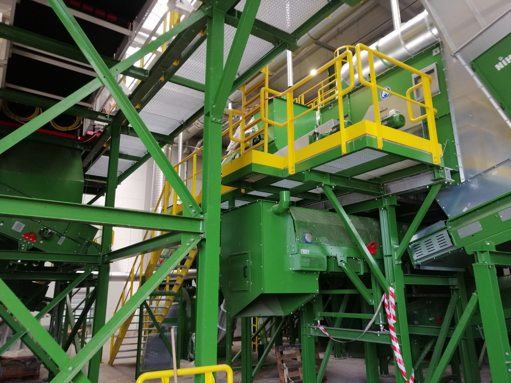 Zielona konstrukcja stalowa hali z żółtymi elementami zabezpieczającymi, widoczne schody i barierki, fragmenty instalacji wentylacyjnej i urządzeń przemysłowych.