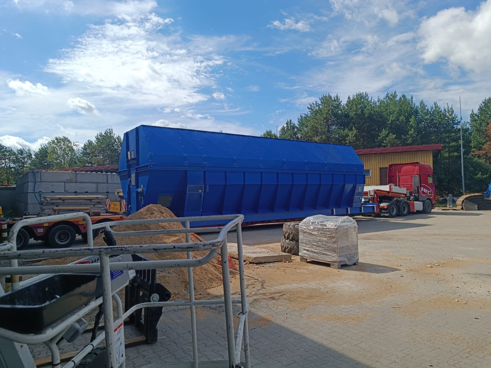 Niebieski kontener przemysłowy na przyczepie, widok z bliska z paletą zafoliowaną obok, na placu z kostki brukowej, w tle budynek i las.