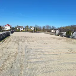 Rozległy plac z kostki brukowej w odcieniach beżu, otoczony zabudową wiejską i błękitnym niebem. Widok z góry ukazuje regularny wzór ułożenia.