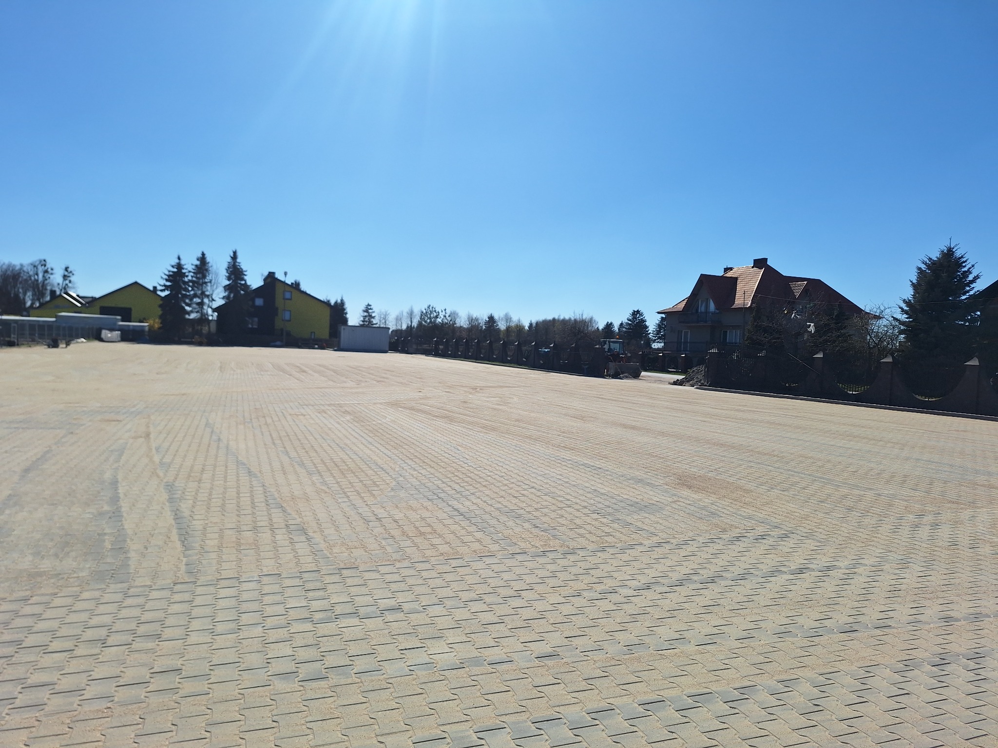 Rozległy plac wyłożony jasnoszarą kostką brukową, otoczony zielenią i budynkami w słoneczny dzień. Widoczne ślady po pojazdach.