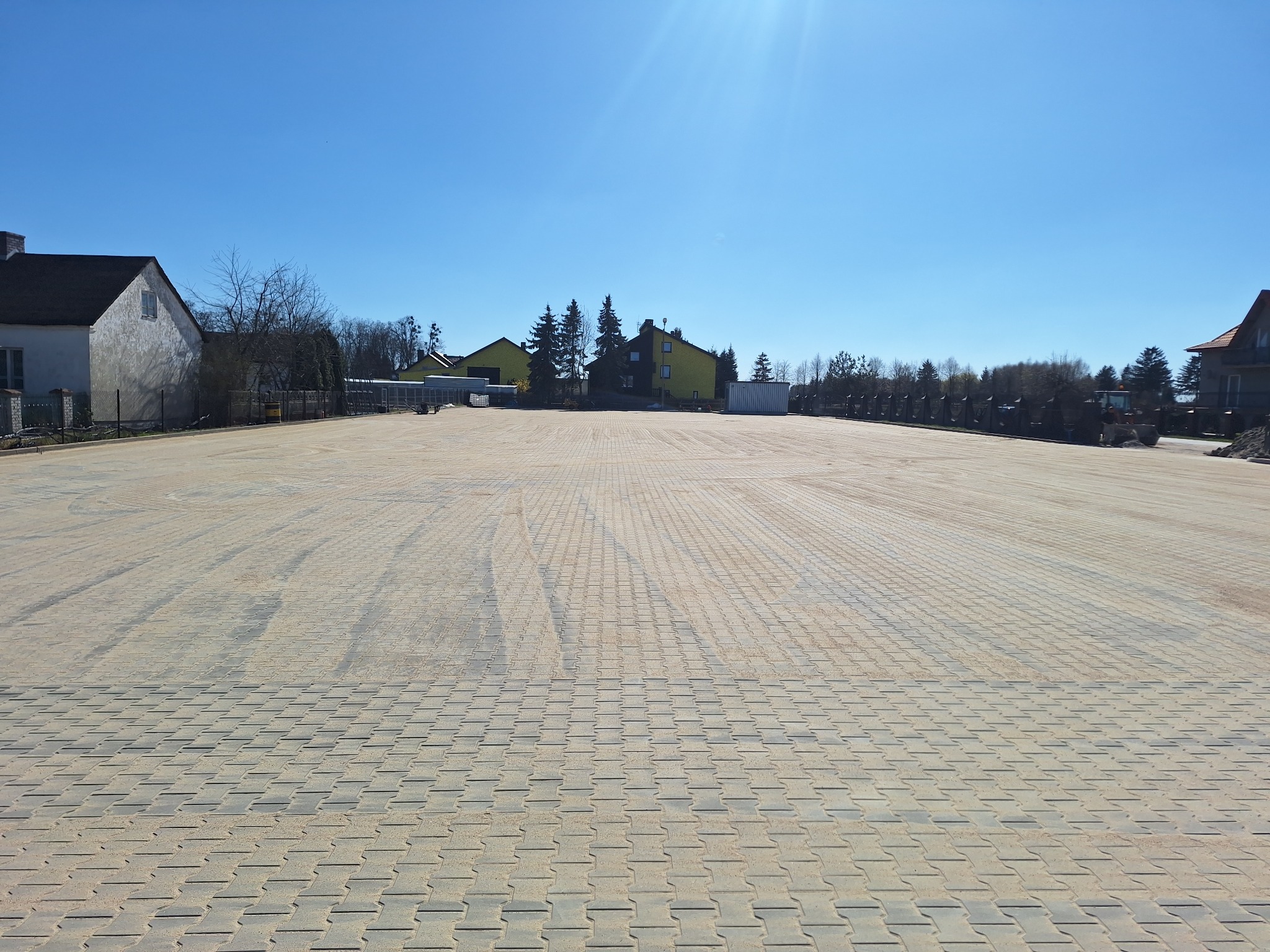 Rozległy, jasnoszary plac wyłożony kostką brukową typu I, z widocznymi śladami po maszynach budowlanych, otoczony zabudową jednorodzinną w słoneczny dzień.