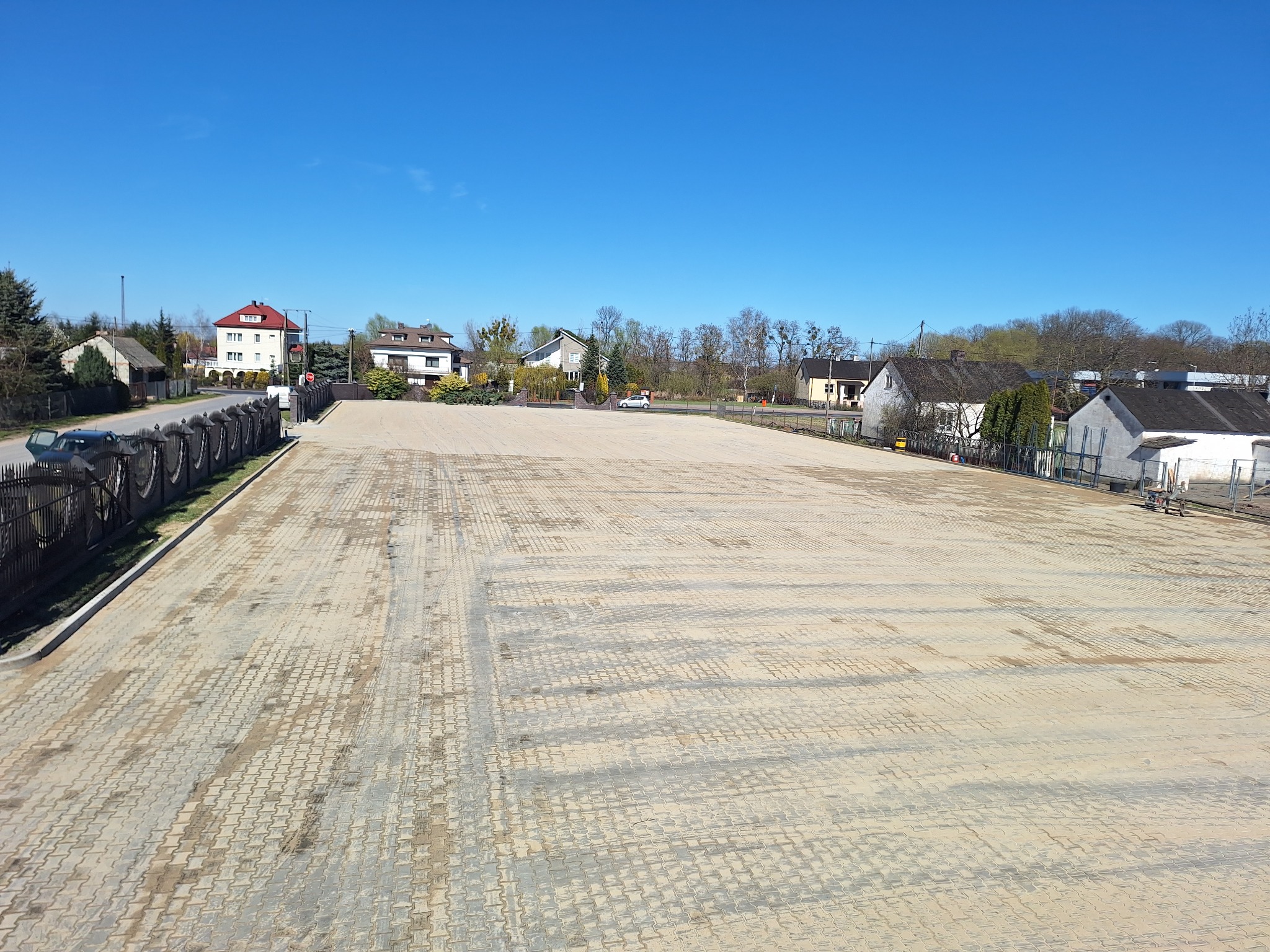 Rozległy plac z kostki brukowej w odcieniach beżu, otoczony zabudową wiejską i błękitnym niebem. Widok z góry ukazuje regularny wzór ułożenia.
