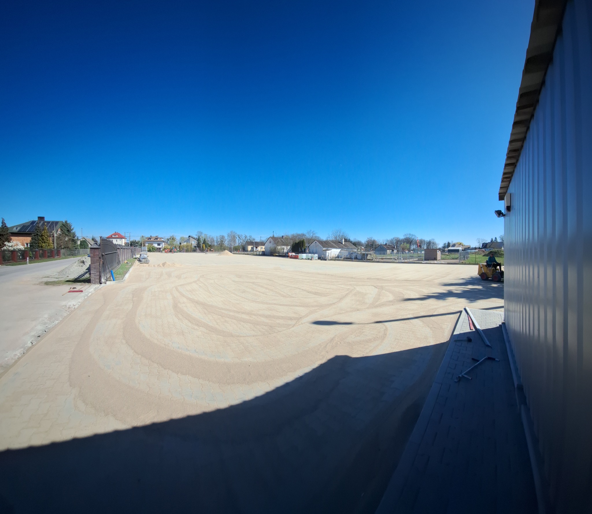 Rozległy teren przygotowany pod kostkę brukową, z widocznymi śladami po maszynach, na tle zabudowań i błękitnego nieba. W tle widać budynki mieszkalne.