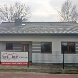 Nowo wybudowany, parterowy dom z szarą dachówką w Skarżysku-Kamiennej, ogrodzony siatką, z widocznym banerem reklamowym firmy budowlanej.