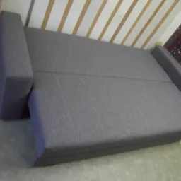 Rozłożona szara sofa z tapicerką o widocznej fakturze, sfotografowana pod kątem, ukazująca jej konstrukcję i mechanizm rozkładania.