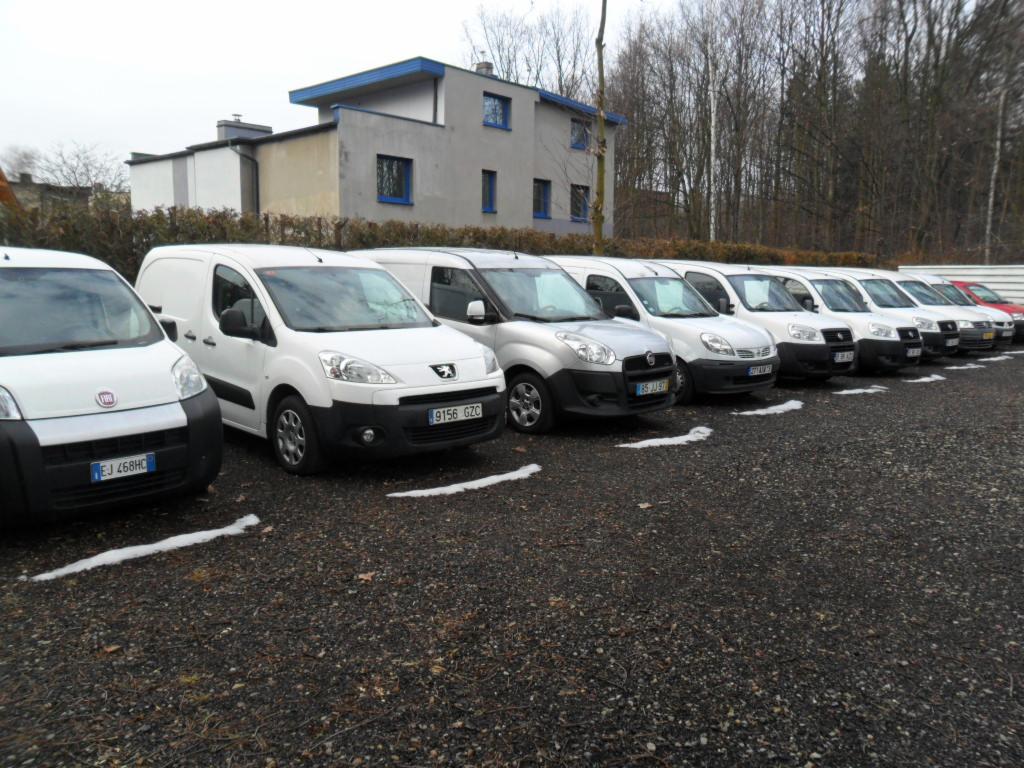 Rząd zaparkowanych aut dostawczych różnych marek, w tym Fiat i Peugeot, stojących na żwirowym parkingu z fragmentami śniegu, na tle budynku i drzew.