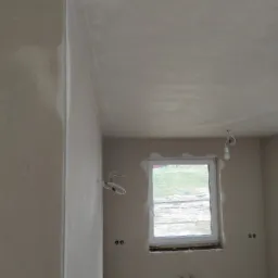 Świeżo otynkowane wnętrze z widocznymi przewodami elektrycznymi i oknem na zewnątrz, sufit i ściany pokryte jasnoszarą, gładką powierzchnią.