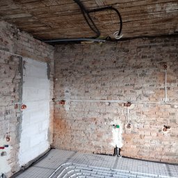 Elektro-Progres - Surowa kuchnia w trakcie remontu: widoczne przewody elektryczne ułożone na ceglanej ścianie, rurki ogrzewania podłogowego na podłodze oraz czarne kable pod sufitem z drewnianych desek.