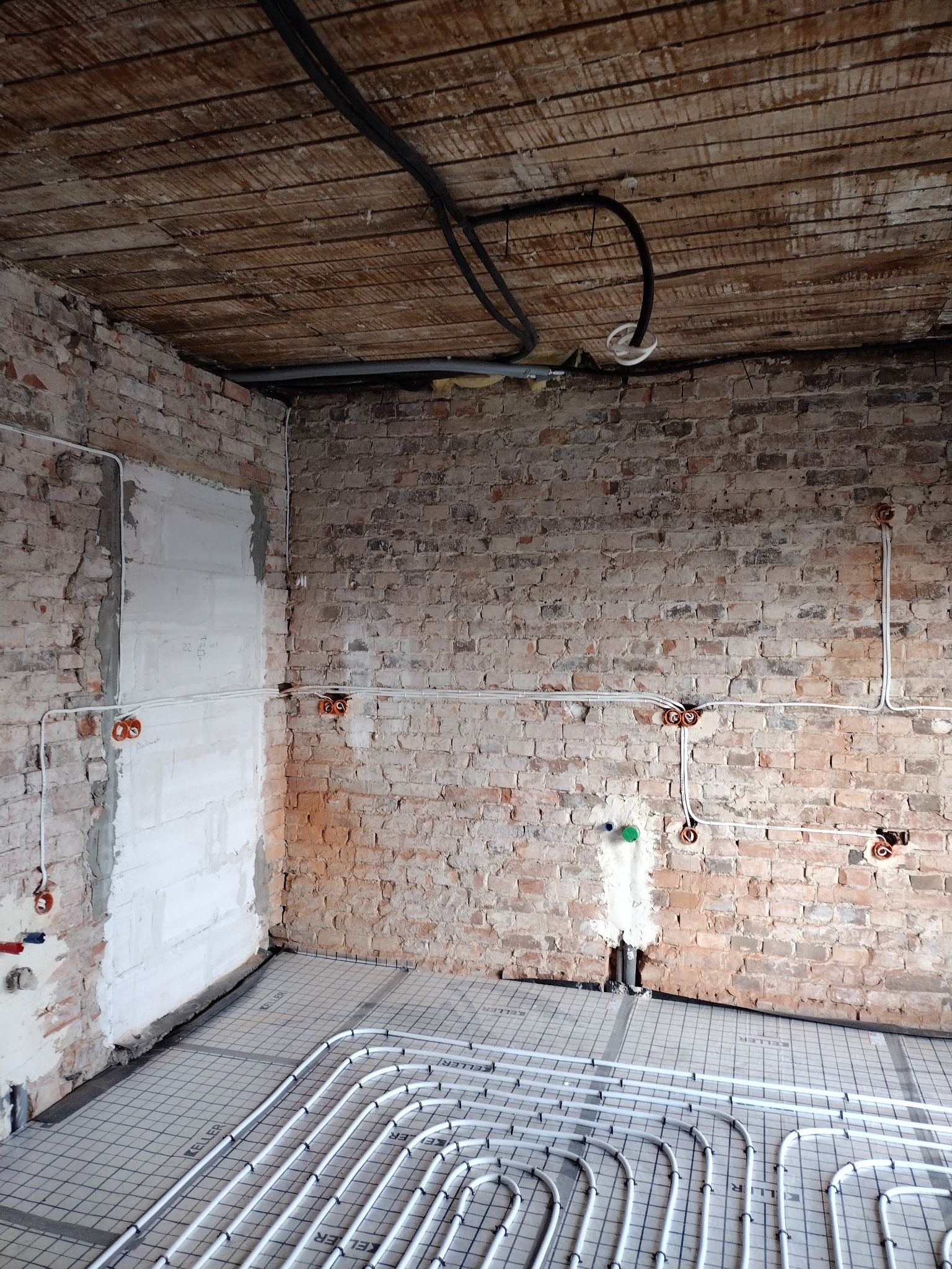 Surowa kuchnia w trakcie remontu: widoczne przewody elektryczne ułożone na ceglanej ścianie, rurki ogrzewania podłogowego na podłodze oraz czarne kable pod sufitem z drewnianych desek.