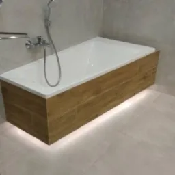 Nowoczesna łazienka z obudowaną wanną z drewnopodobnych płytek i subtelnym podświetleniem LED u dołu, szare płytki na ścianach i podłodze, chromowana armatura.