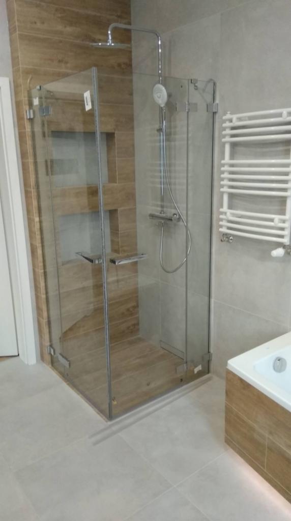 Kabina prysznicowa ze szklanymi drzwiami z chromowanymi zawiasami, wykończona brązowymi płytkami imitującymi drewno, z chromowaną armaturą i grzejnikiem drabinkowym w nowoczesnej łazience.