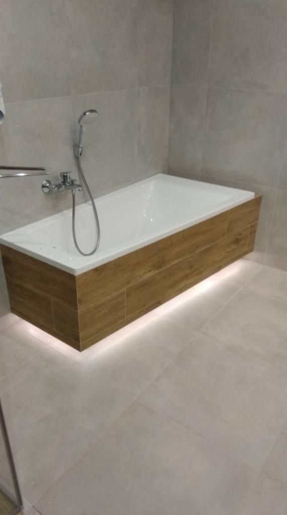 Nowoczesna łazienka z obudowaną wanną z drewnopodobnych płytek i subtelnym podświetleniem LED u dołu, szare płytki na ścianach i podłodze, chromowana armatura.
