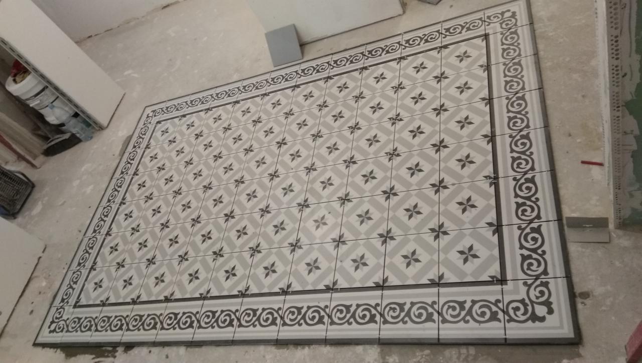 Ułożona podłoga z płytek ceramicznych w geometryczny wzór z dekoracyjnym obramowaniem, widoczne resztki zaprawy i narzędzie glazurnicze.