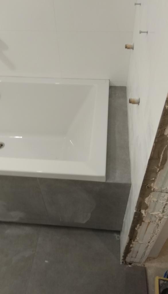 Biała wanna obudowana szarymi płytkami, widoczne fragmenty ściany w trakcie wykańczania z wystającymi elementami montażowymi.