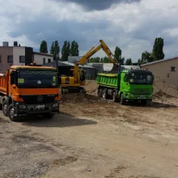 Żółta koparka Komatsu ładuje urobek do zielonej wywrotki Mercedes Actros, pomarańczowa wywrotka stoi obok, w tle budynki i hałdy ziemi.