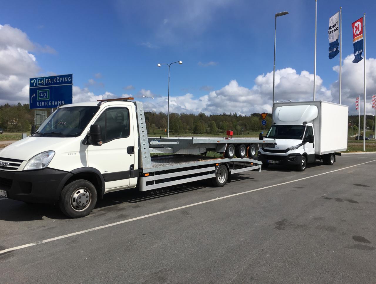 Biała laweta Iveco z platformą i biały bus Iveco na parkingu, w tle drogowskaz na Falköping i Ulricehamn oraz flagi firmowe.