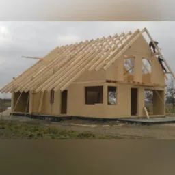 Wznoszenie drewnianej konstrukcji domu z widoczną więźbą dachową i pracownikami na rusztowaniu.