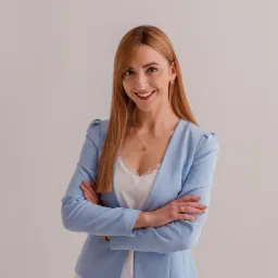 Portret uśmiechniętej kobiety w błękitnym żakiecie na jasnym tle, z rękami skrzyżowanymi na piersiach, widoczne detale biżuterii: kolczyki i naszyjnik z zawieszką w kształcie serca.