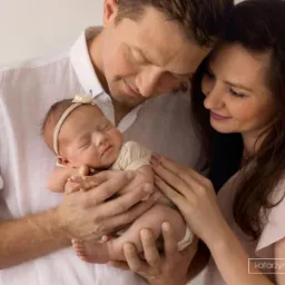 Rodzice przytulają śpiące nowonarodzone dziecko z opaską na głowie, delikatne światło, ciepła kolorystyka.