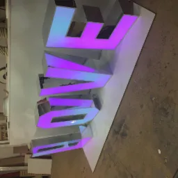 Podświetlany, trójwymiarowy napis LOVE z tworzywa sztucznego w odcieniach fioletu i błękitu, leżący na płycie w warsztacie stolarskim, widoczne narzędzia i elementy wyposażenia.