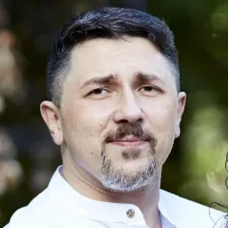 Portret mężczyzny z szpakowatą brodą w białej koszuli na rozmytym tle. Ujęcie od przodu, skupione na twarzy i wyrazie.