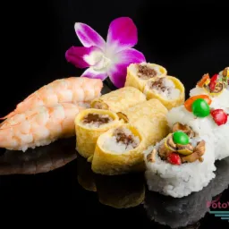 Kompozycja sushi z krewetkami, naleśnikami i kolorowymi dodatkami, ozdobiona fioletowym kwiatem na czarnym, błyszczącym tle.