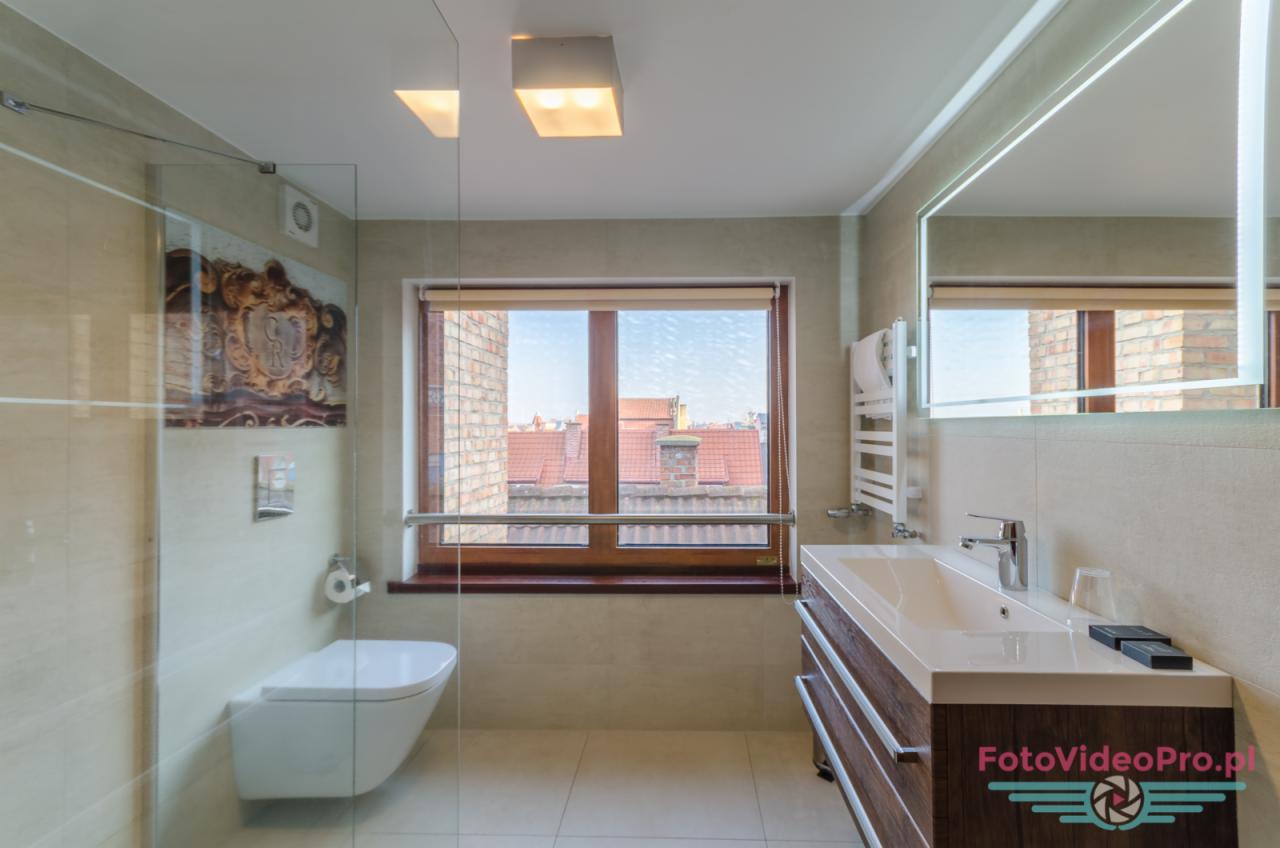 Nowoczesna łazienka z przeszkloną kabiną prysznicową, wiszącą toaletą i umywalką z szafką z drewnopodobnym fornirem, widok na dachy budynków przez okno.