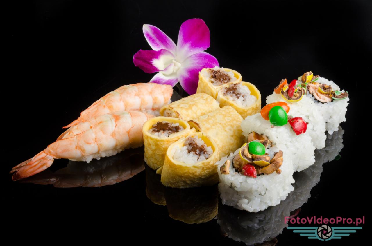 Kompozycja sushi z krewetkami, naleśnikami i kolorowymi dodatkami, ozdobiona fioletowym kwiatem na czarnym, błyszczącym tle.