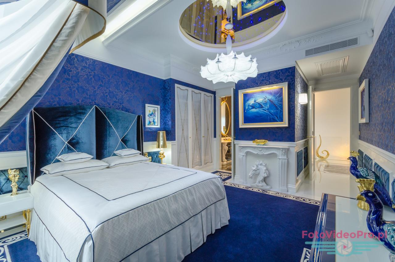 Luksusowa sypialnia w odcieniach błękitu i bieli z eleganckim łóżkiem, dekoracyjnym kominkiem z rzeźbą konia i kryształowym żyrandolem.