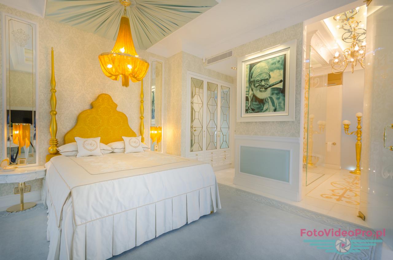 Luksusowa sypialnia z baldachimem i złotymi elementami, widok na łazienkę z kryształowym żyrandolem, na ścianie portret palącego cygaro mężczyzny.