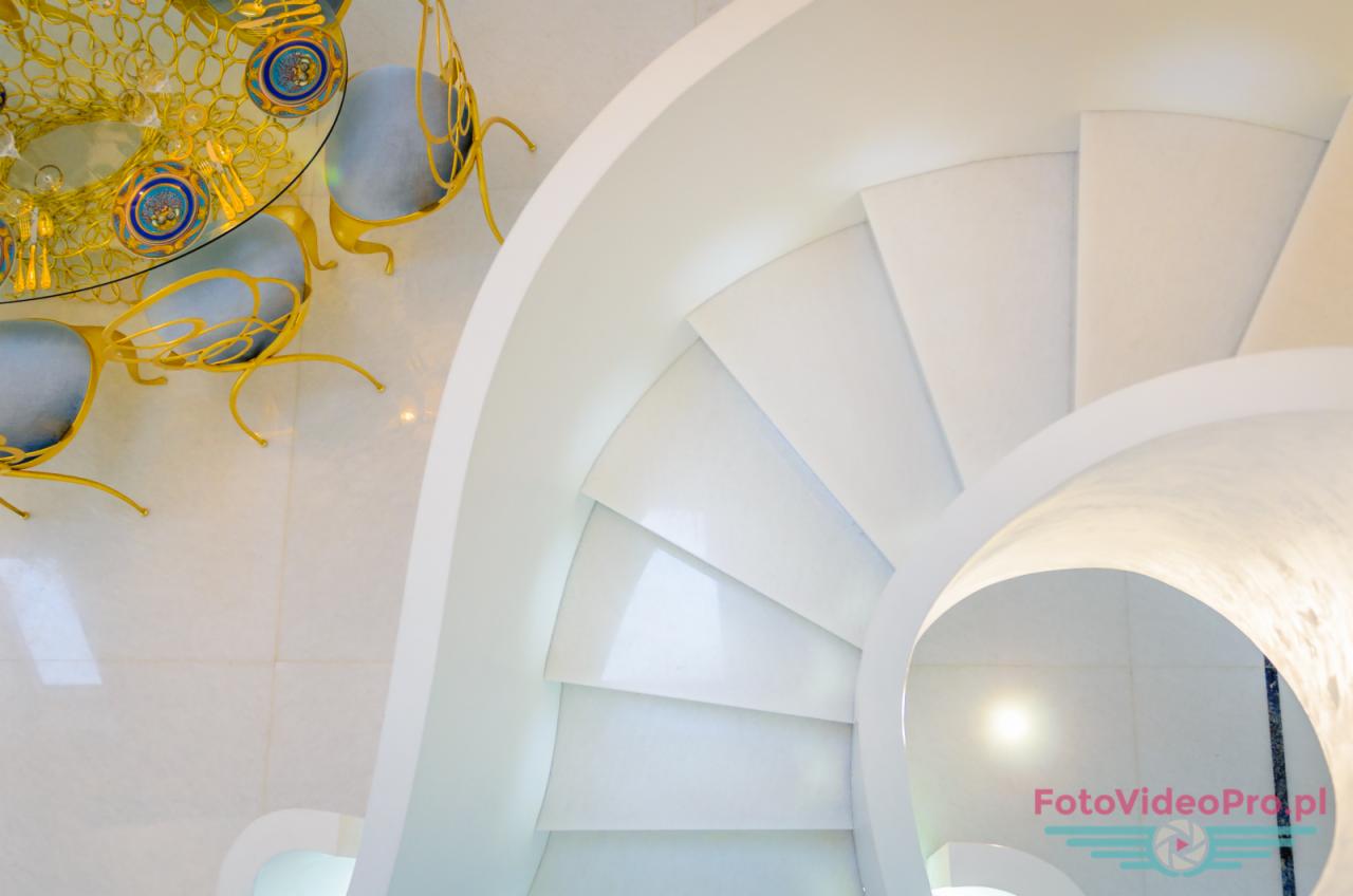 Białe spiralne schody z widokiem na stół z krzesłami o złotych nogach i niebieskich siedziskach