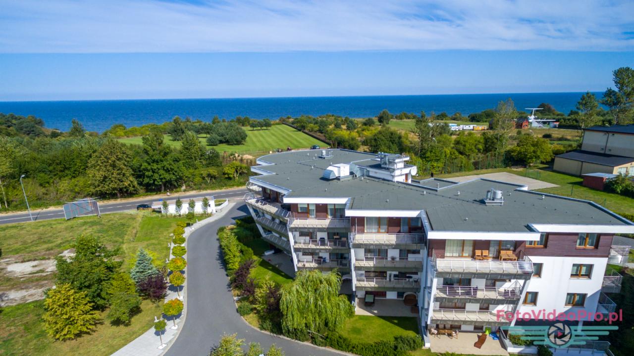 Widok z lotu ptaka na budynek apartamentowy z balkonami, położony blisko morza i terenów zielonych, z widocznym helikopterem w tle.