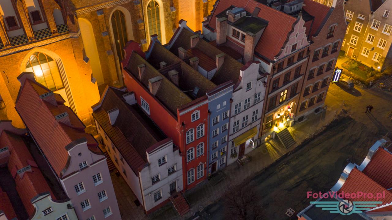 Widok z lotu ptaka na kolorowe kamienice w Gdańsku o zmierzchu, z widocznym oświetlonym kościołem w tle. Dachówki w odcieniach czerwieni i pomarańczy, oraz logo w prawym dolnym rogu.