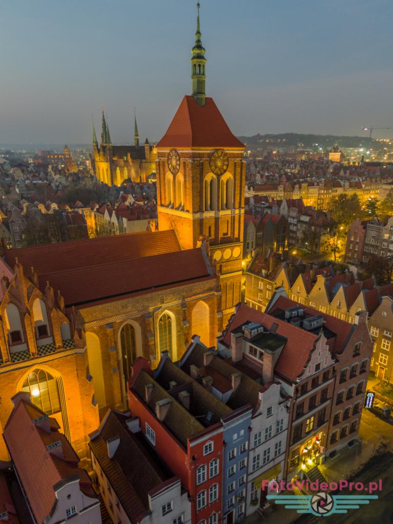 Panorama Gdańska nocą z lotu ptaka, oświetlony kościół Mariacki z charakterystyczną wieżą zegarową na pierwszym planie, w tle inne budynki Starego Miasta.
