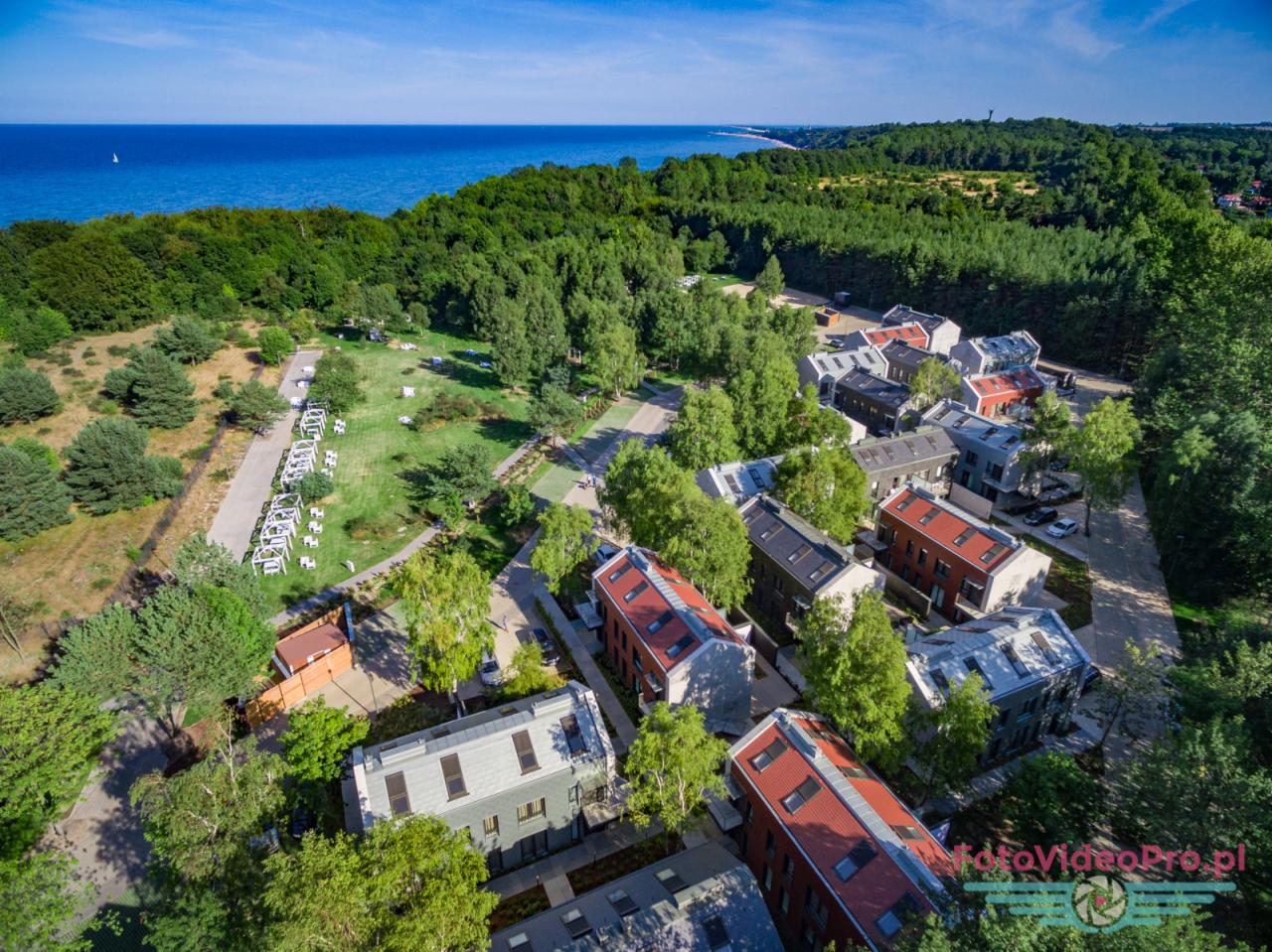 Widok z lotu ptaka na nowoczesne osiedle domków jednorodzinnych z czerwonymi i szarymi dachami, położone w otoczeniu zieleni, z widokiem na morze i plażę w oddali; na trawniku przygotowania...