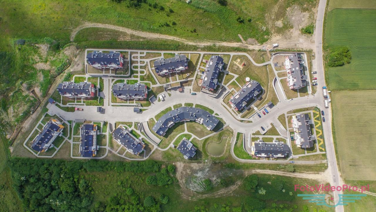 Zdjęcie z drona przedstawiające osiedle nowych budynków wielorodzinnych z placem zabaw, drogami dojazdowymi i zielenią, widziane z góry.