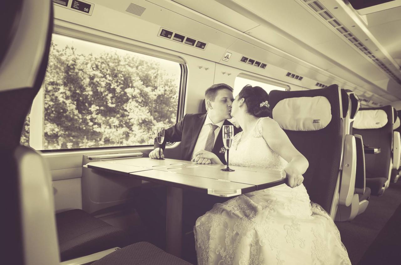Nowożeńcy w pociągu w Warszawie, delikatny pocałunek przy stoliku z kieliszkami szampana, widok za oknem na zieleń.