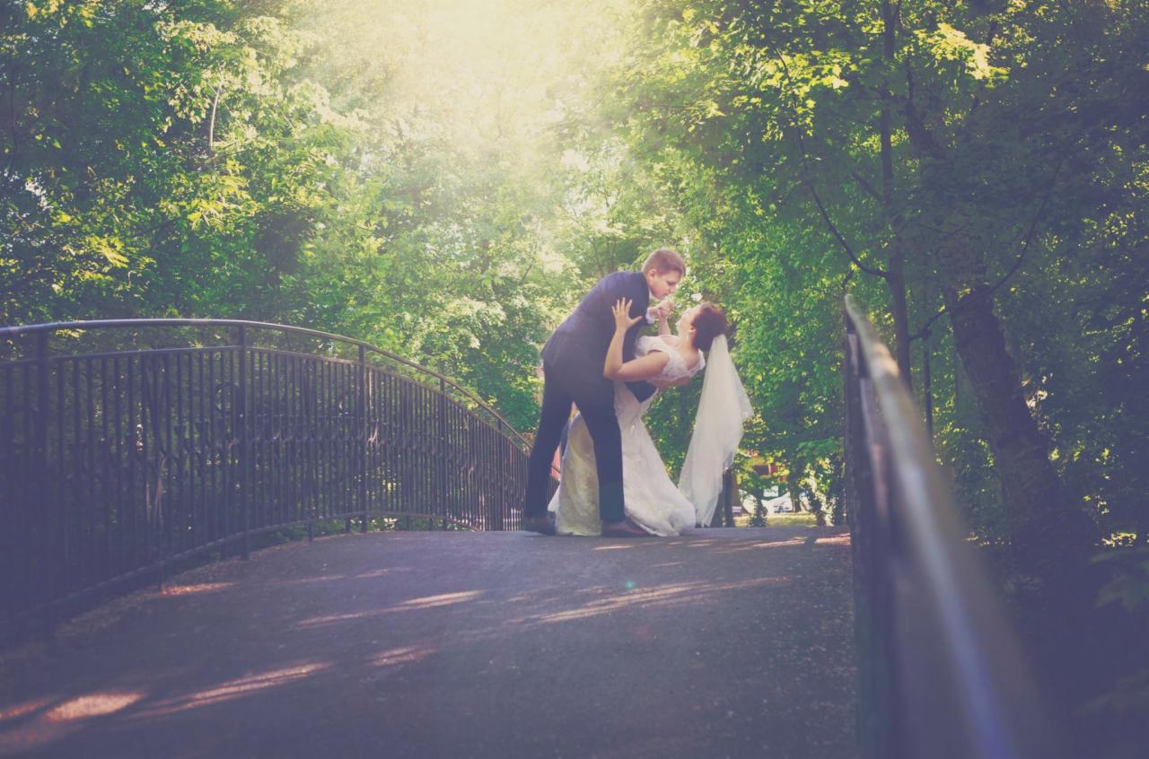 Para młoda w objęciach na moście w parku, on w ciemnym garniturze, ona w białej sukni ślubnej z welonem, otoczeni zielenią drzew, w tle delikatne światło słoneczne.