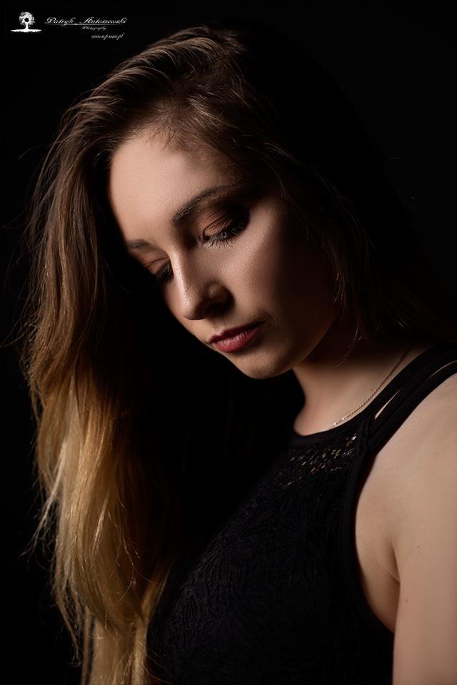 Portret kobiety z długimi, blond włosami w czarnej koronkowej bluzce, spuszczony wzrok, ciemne tło, oświetlenie podkreślające kontury twarzy.