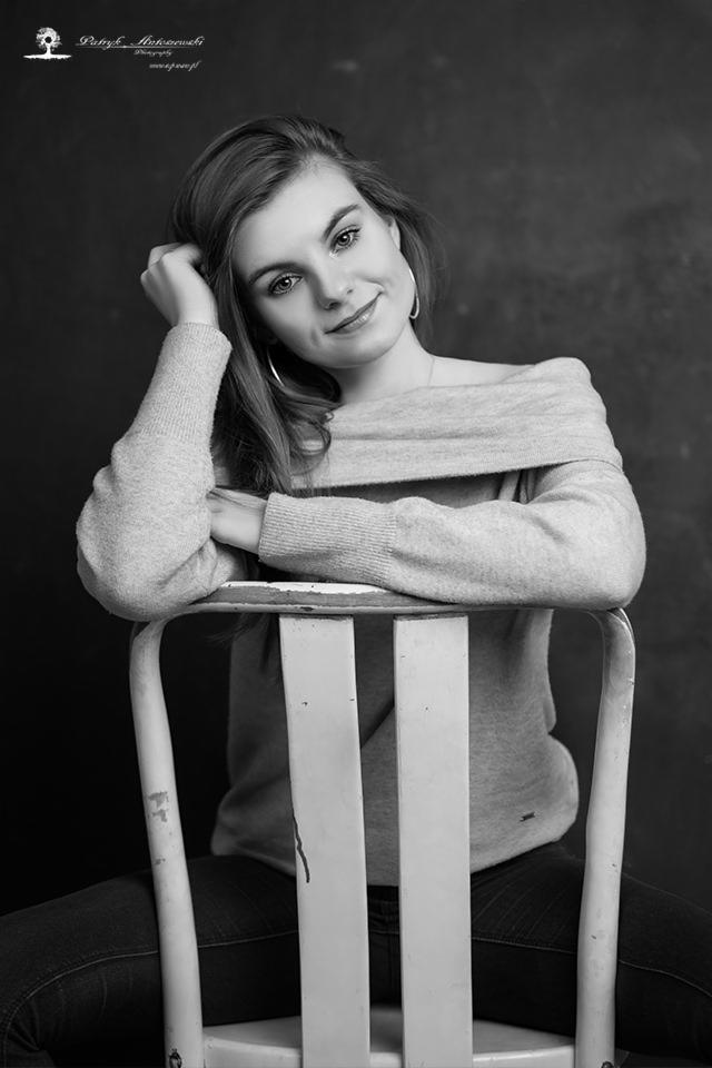 Portret młodej kobiety w swetrze z odkrytymi ramionami, opierającej się o białe krzesło na ciemnym tle, wykonany w czerni i bieli z delikatnym uśmiechem.