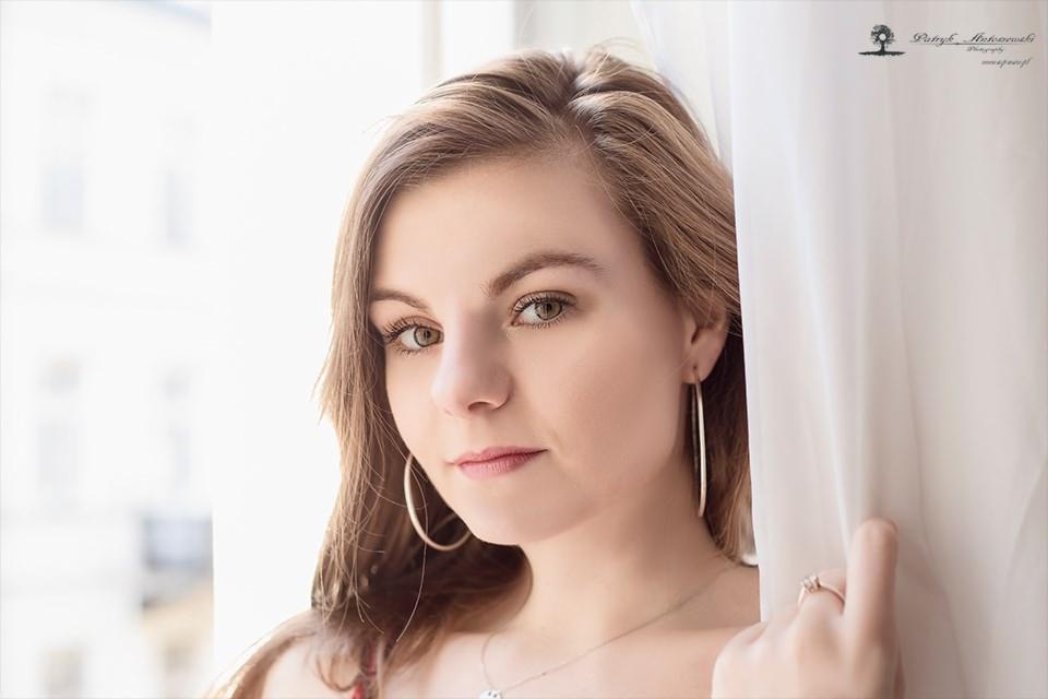 Portret młodej kobiety z długimi, prostymi włosami, dużymi kolczykami kołami i delikatnym makijażem, pozującej przy oknie z jasną firanką trzymaną w dłoni z pierścionkiem.