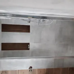 Wykończona łazienka z szarymi płytkami imitującymi beton, drewnopodobnymi panelami na ścianie, wnękami w ścianie pod prysznicem i chromowaną armaturą.