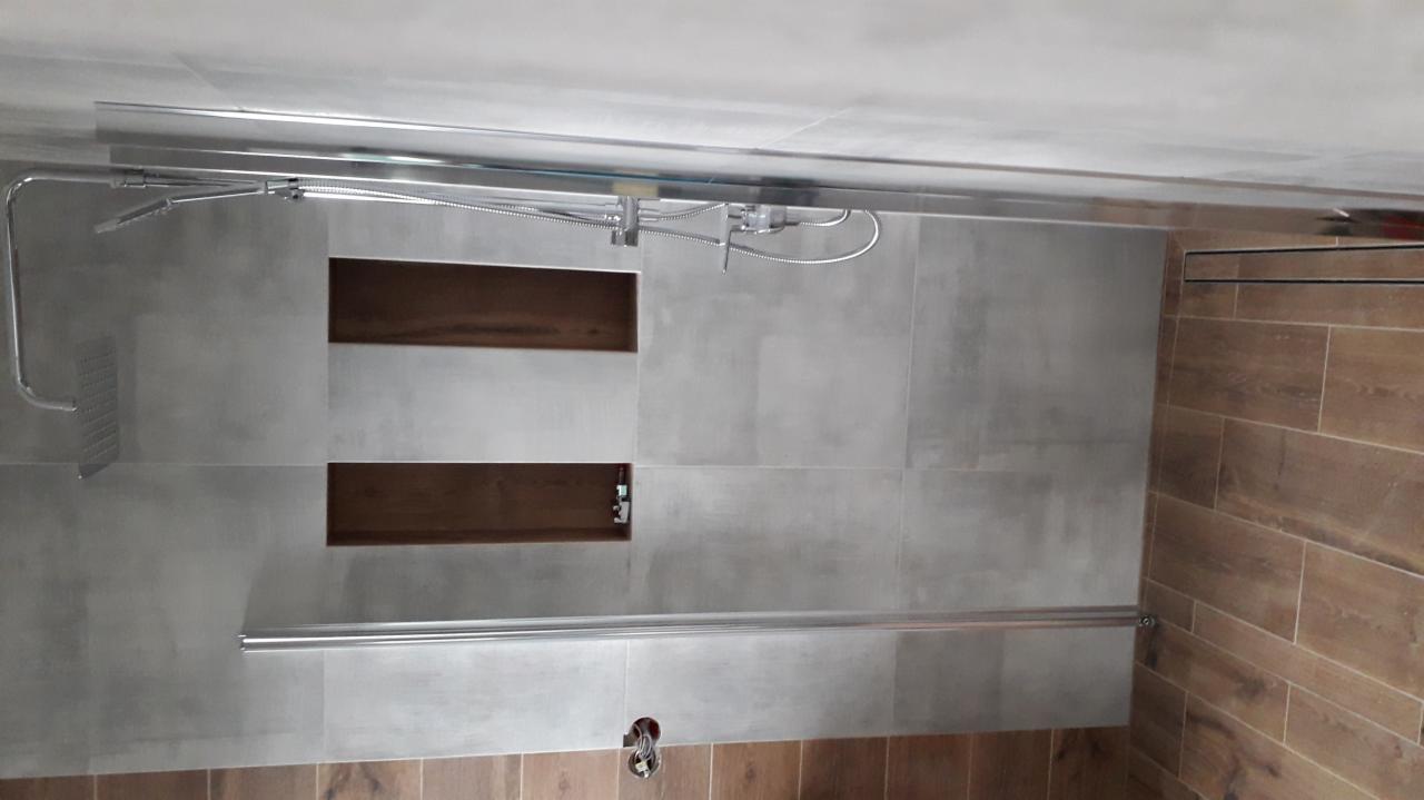 Wykończona łazienka z szarymi płytkami imitującymi beton, drewnopodobnymi panelami na ścianie, wnękami w ścianie pod prysznicem i chromowaną armaturą.