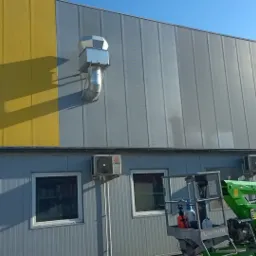 Wysoka hala przemysłowa z żółto-szarą elewacją z blachy trapezowej, widoczny podnośnik koszowy z operatorem przygotowany do pracy na wysokości, klimatyzatory na ścianie.