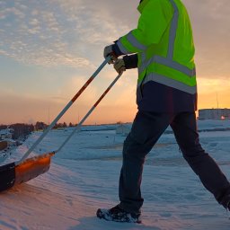 Zieleń i Drewno - Pracownik w odblaskowej kurtce usuwa śnieg z dachu płaską, szeroką łopatą na długim uchwycie, na tle zachodzącego słońca i zabudowań miejskich.