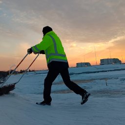 Zieleń i Drewno - Pracownik w jaskrawej kurtce odblaskowej odśnieża dach płaski za pomocą specjalnej łopaty dwuręcznej na tle wschodzącego słońca.