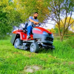 Mężczyzna z brodą na czerwonej kosiarce samojezdnej tnący wysoką trawę na zielonym trawniku, w tle drzewa i drewniany płot.