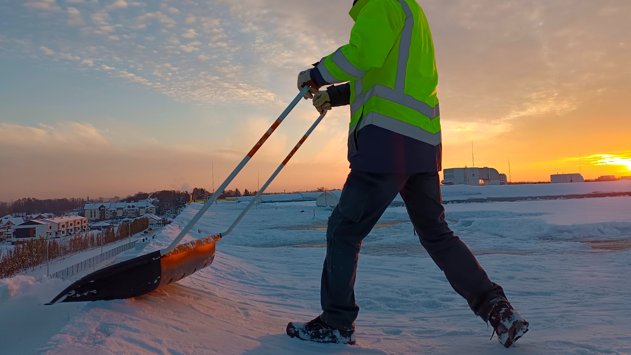 Pracownik w odblaskowej kurtce usuwa śnieg z dachu płaską, szeroką łopatą na długim uchwycie, na tle zachodzącego słońca i zabudowań miejskich.