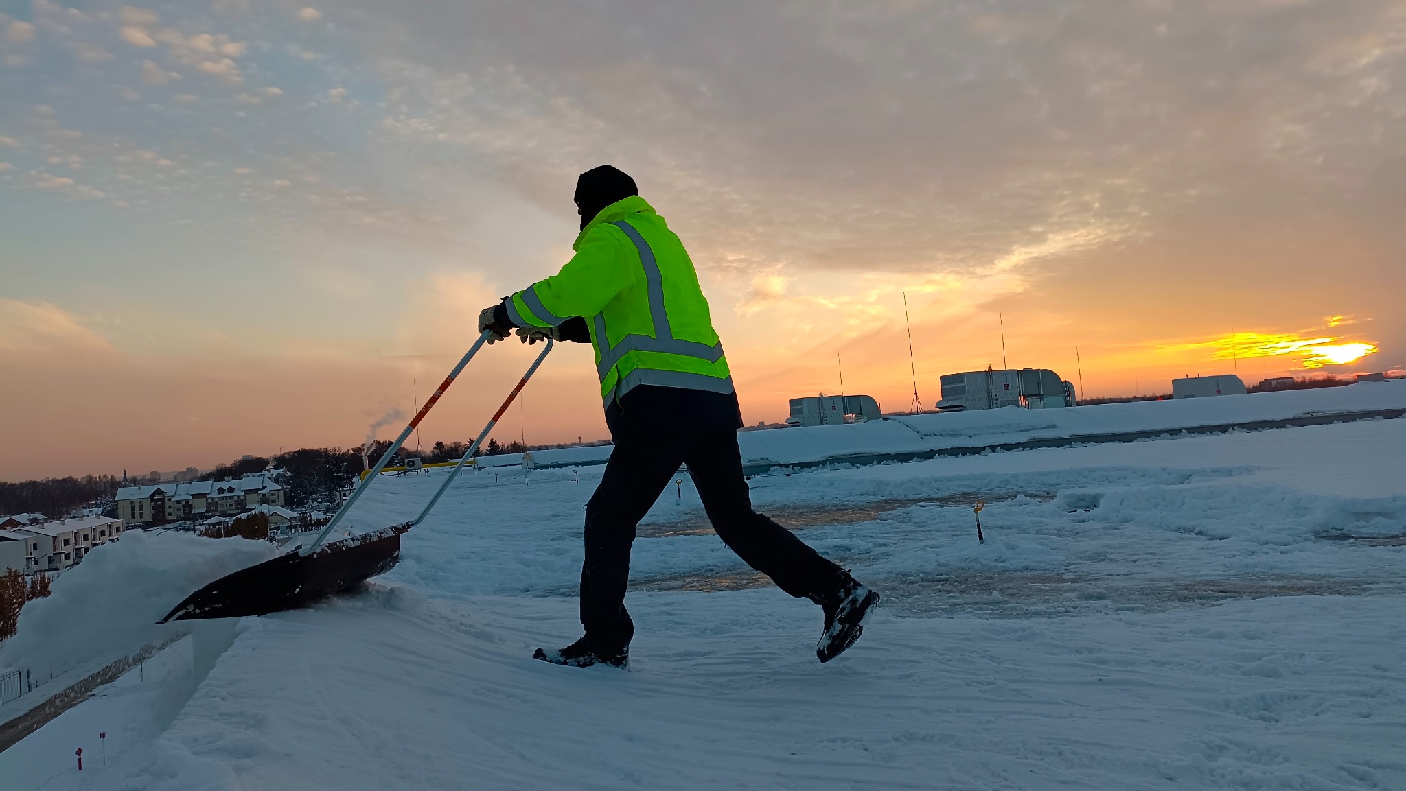 Pracownik w jaskrawej kurtce odblaskowej odśnieża dach płaski za pomocą specjalnej łopaty dwuręcznej na tle wschodzącego słońca.